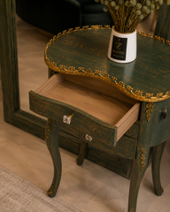 A dark green distressed wooden nightstand with ornate gold trim and an open drawer, styled with a white vase of dried flowers on top.