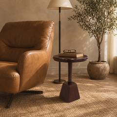 A mid-century modern side table with a dark wood round top and geometric pedestal base, styled in a warm living room next to a brown leather armchair and a floor lamp.