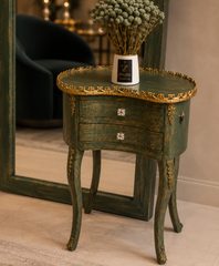 A distressed dark green wooden side table with ornate gold trim and curved cabriole-style legs, styled with a white vase of dried greenery on top, set in an indoor room with a mirror in the background.