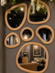 A set of organic-shaped wooden mirrors with light natural wood frames, arranged on a dark gray wall. The mirrors reflect a furnished interior with a sofa, side table, and framed artwork, while also showing a dark ceiling with track lighting and hanging greenery and white flowers.