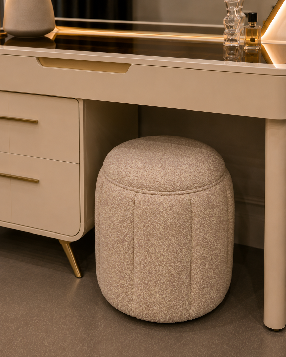 A cylindrical upholstered ottoman stool in a light beige bouclé fabric, positioned underneath a cream-colored vanity desk with gold accents.