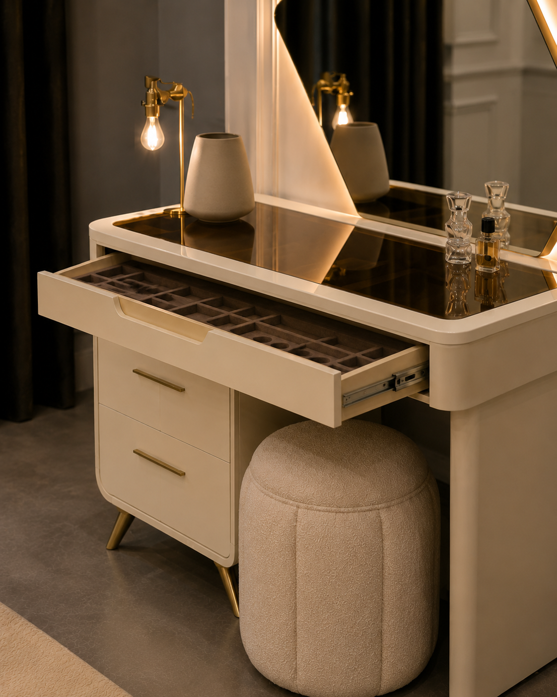 A cream-colored vanity table with gold-tone hardware, featuring an open drawer, a round upholstered ottoman, and a large illuminated mirror. The vanity is styled with a vase, perfume bottles, and a gold filament lamp in a modern, dimly lit interior.