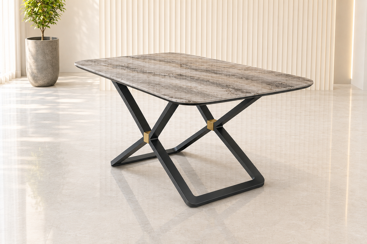 A modern rectangular dining table with a gray and white marbled top and a black metal X-shaped base with gold accents, set in a bright, modern room with a potted plant in the background.