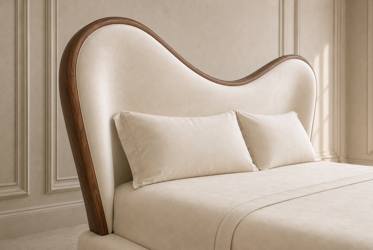 A close-up of a modern upholstered bed headboard with a curved, wave-like silhouette, finished in cream fabric with a dark brown wood frame, shown with matching cream bedding and pillows in a softly lit bedroom.
