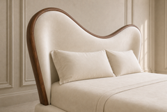 A close-up of a modern upholstered bed headboard with a curved, wave-like silhouette, finished in cream fabric with a dark brown wood frame, shown with matching cream bedding and pillows in a softly lit bedroom.