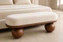 A modern upholstered bench with a cream fabric cushion and rounded wooden legs, positioned in front of a matching cream upholstered bed in a bright, neutral-toned room.