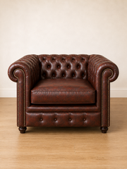 A classic Chesterfield armchair in dark brown leather with tufted button detailing, rolled arms, and nailhead trim, set against a neutral beige wall and light wood floor.