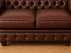 A close-up of a dark brown leather Chesterfield sofa, showing the tufted backrest, two seat cushions, and the matching tufted base with dark wooden legs on a light wood floor.