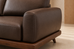 Close-up of a dark brown leather sofa armrest with a wooden base and leg, shown in a modern interior setting.
