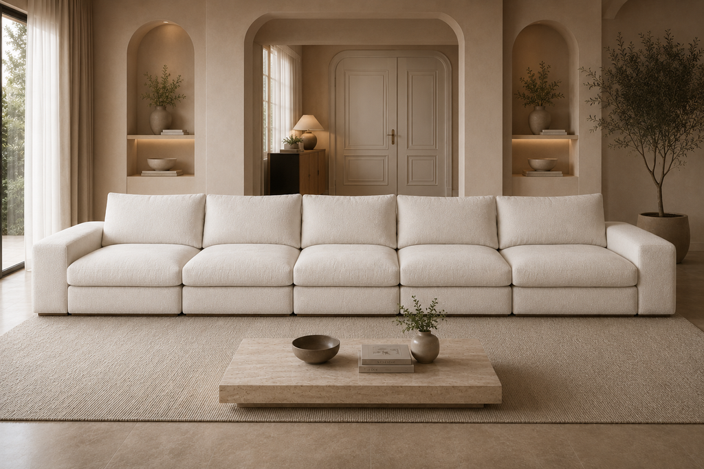 A modern, low-profile modular sectional sofa upholstered in a warm off-white fabric, set in a beautifully styled, minimalist living room with beige walls, a wooden coffee table, and built-in arched shelves.