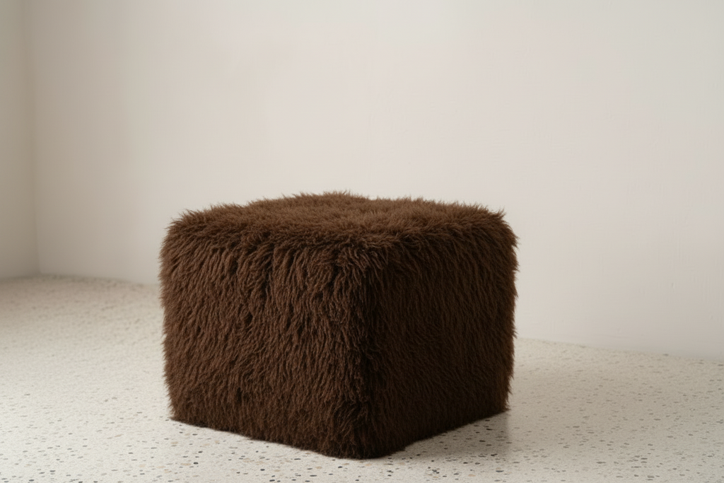 Brown fluffy cube-shaped ottoman on a light carpet with a white wall background