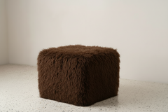 Brown fluffy cube-shaped ottoman on a light carpet with a white wall background