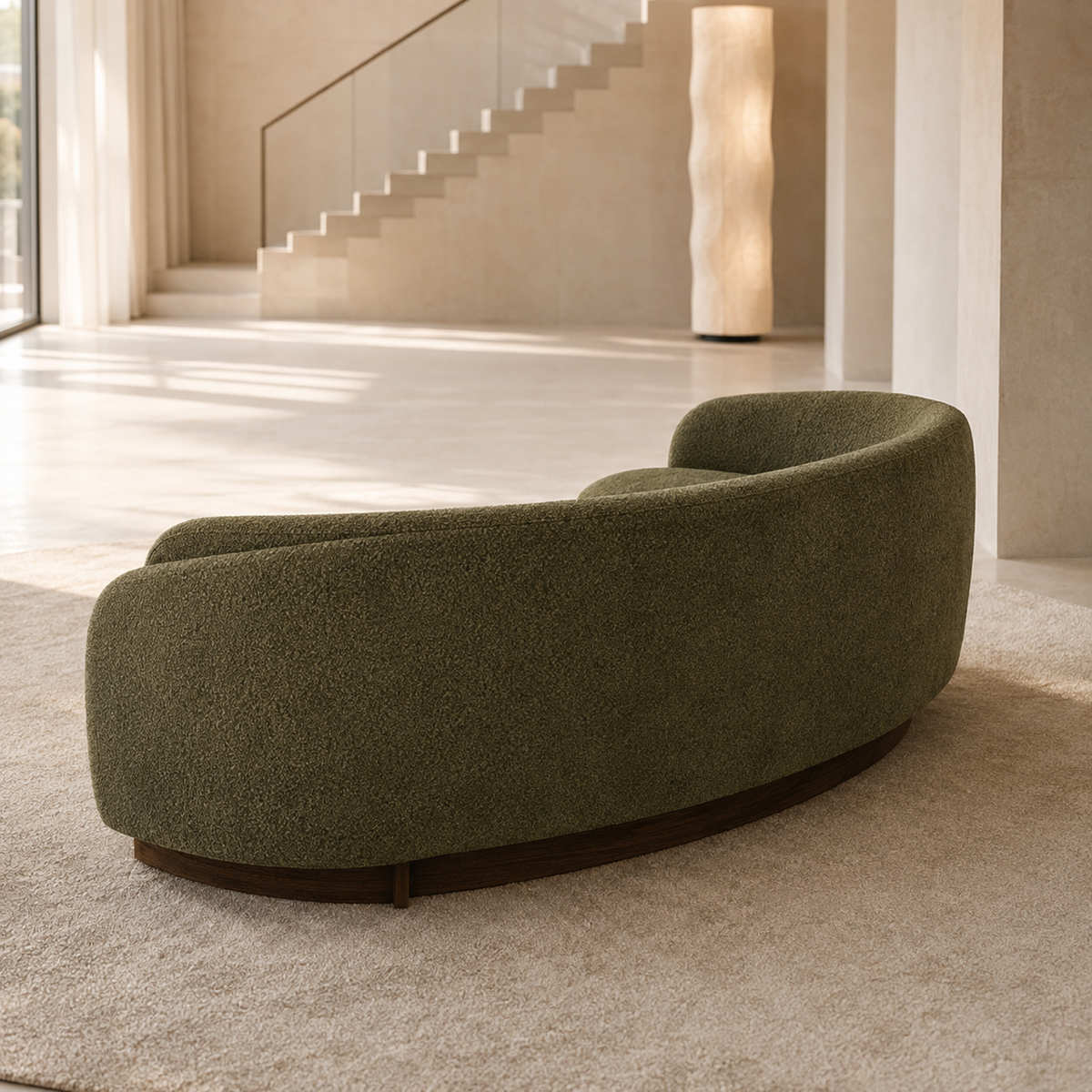 A curved, low-profile upholstered sofa in textured olive green fabric with a dark wood base, placed on a light beige rug in a modern minimalist interior with a staircase in the background.