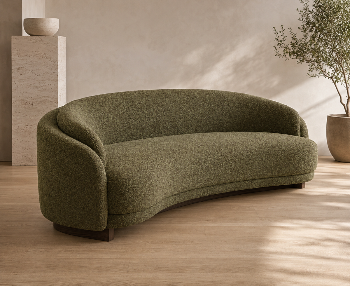 A curved, low-profile sofa upholstered in textured olive green bouclé fabric, set in a minimalist room with light wood floors, a beige wall, and a potted plant.