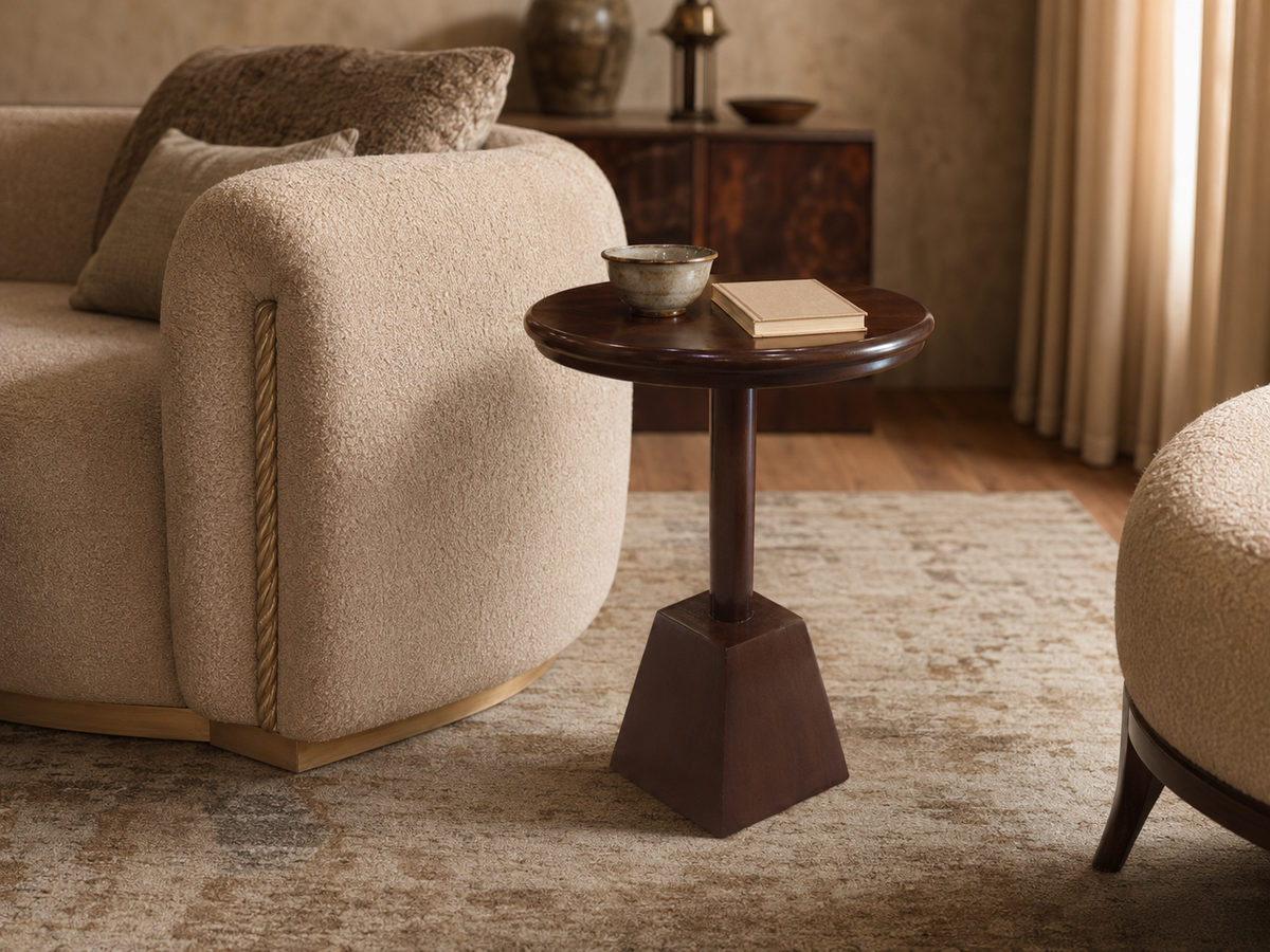 A dark brown wooden pedestal side table with a geometric base, styled in a cozy living room next to a beige bouclé armchair.