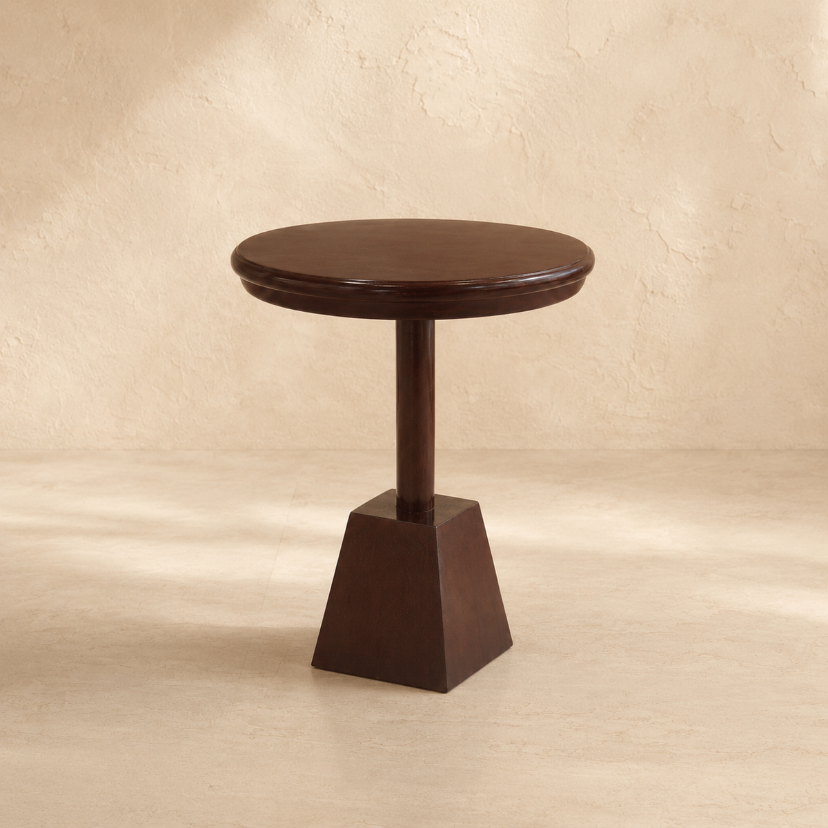 A dark brown wooden pedestal side table with a round top and a geometric square base, set against a textured beige wall and floor.