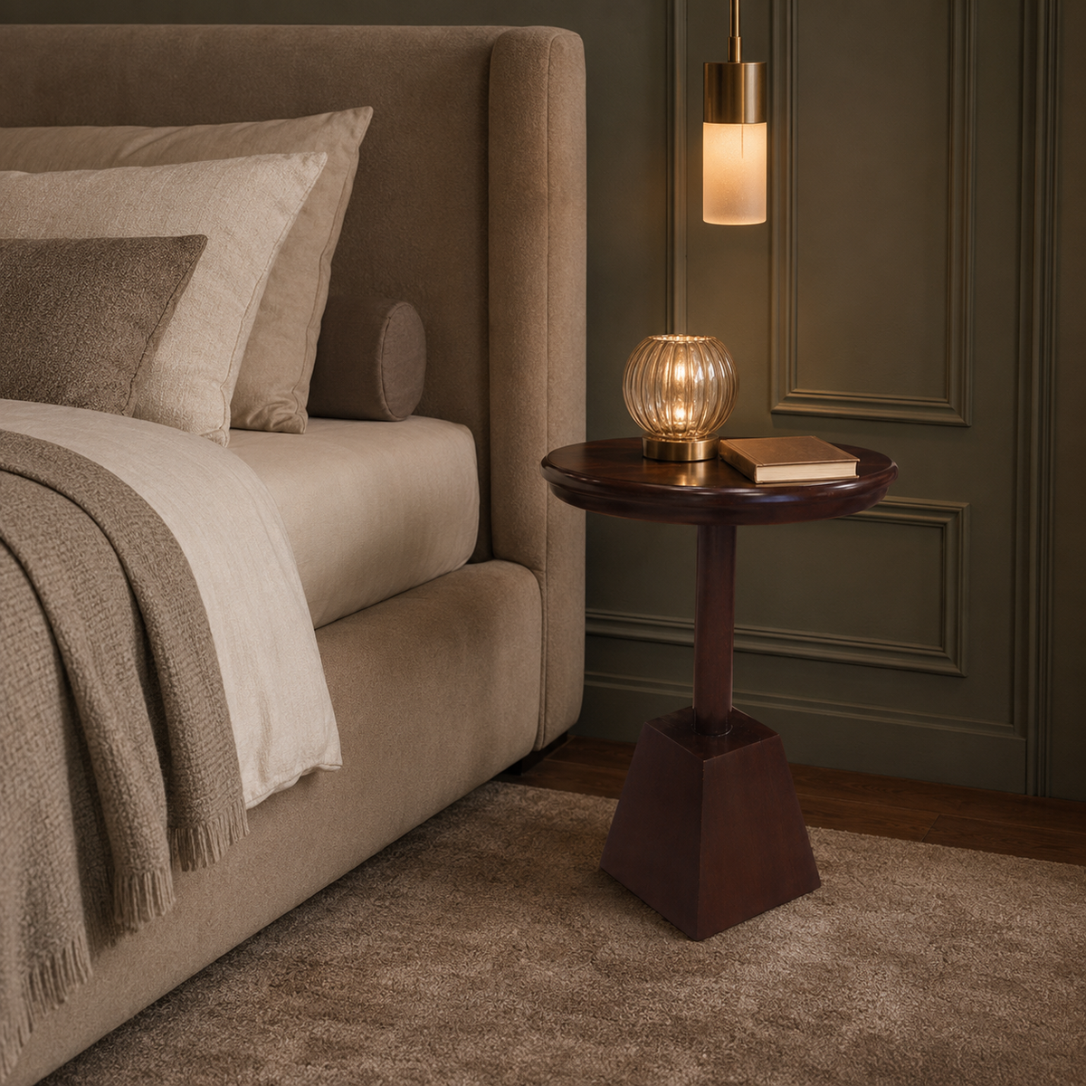A styled bedroom corner showing part of a bed with neutral bedding, a dark wood side table, a lit ribbed glass candle holder, a hanging lamp, and an olive paneled wall.